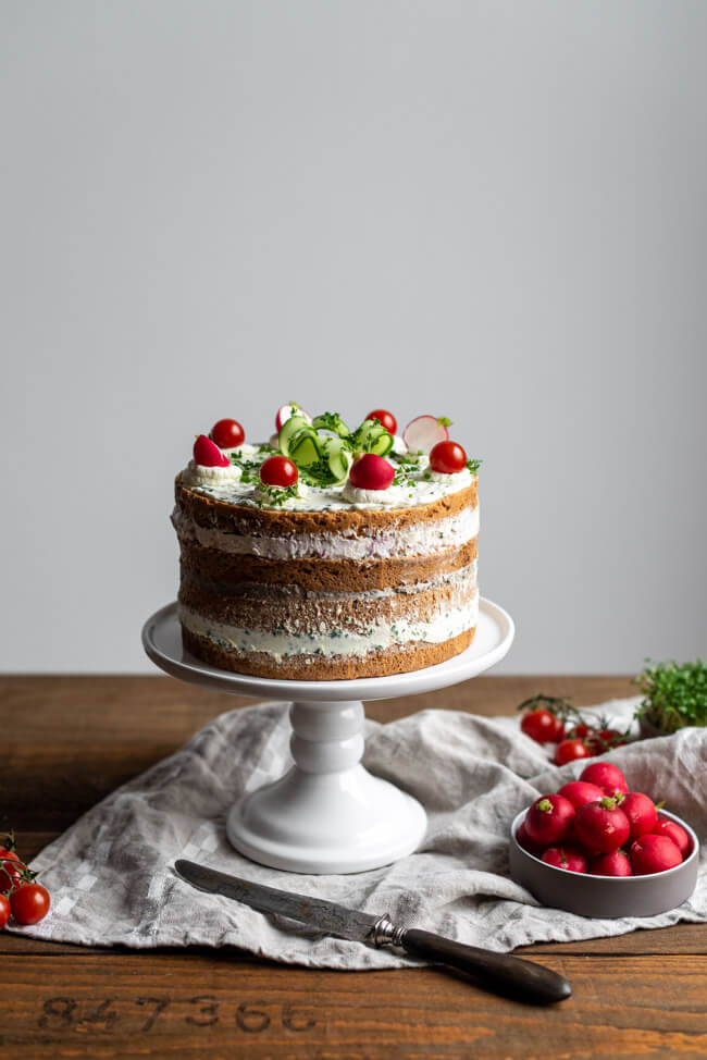 Brotzeittorte mit Pfälzer Leberwurst, Schnittlauchfrischkäsecreme und Radieschen-Frischkäsecreme. Dekoriert mit Meerrettichsahne, Cherrytomaten und Gurken auf weißer Tortenplatte.
