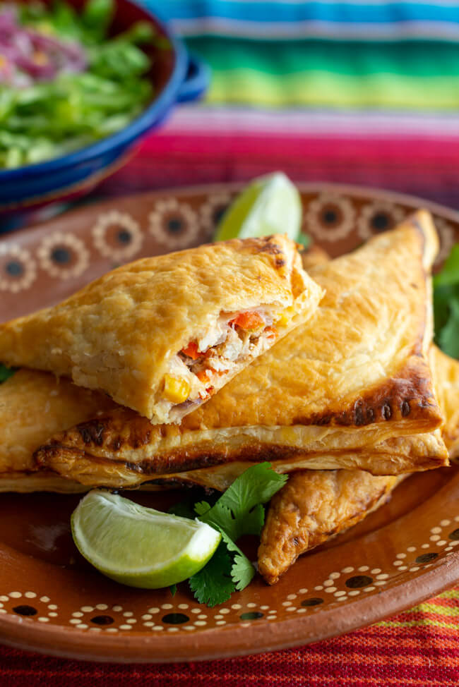Aufgeschnittene TexMex Blätterteigtaschen mit Tabasco und Limette auf rustikalem Tonteller. Dazu eine bunte Keramikschale mit Salat.