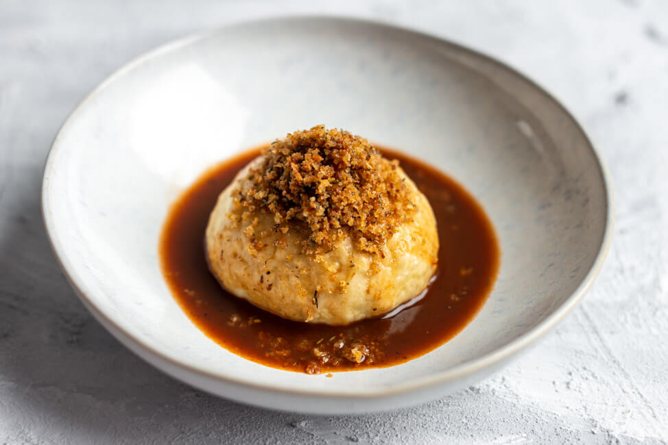 Herzhafter Germknödel mit 'zupfter Goiß, gebratener Gemüsesauce und Thymianbröseln auf weißem Keramikteller und hellem Untergrund.