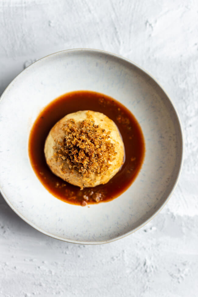 Herzhafter Germknödel mit 'zupfter Goiß, gebratener Gemüsesauce und Thymianbröseln auf weißem Keramikteller und hellem Untergrund.