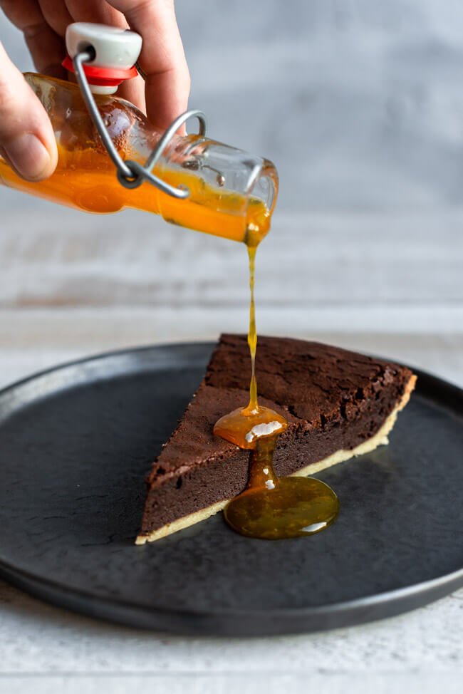 Ein Stück Schokoladentarte auf schwarzem Keramikteller wird mit Mandarinen-Sirup begossen.