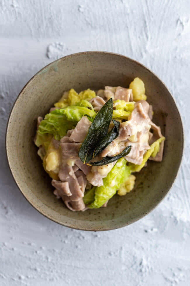 Pizzoccheri Valtellinesi - Pasta aus Buchweizenmehl mit Wirsing, Kartoffeln, Salbei, Knoblauch, brauner Butter und Käse - aus Kitchen Impossible in grauem Keramikteller auf hellem Untergrund.