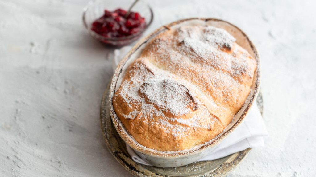 Salzburger Nockerl mit Puderzucker bestäubt aus Kitchen Impossible in Auflaufform auf hellem Untergrund mit Schale Cranberry Kompott.