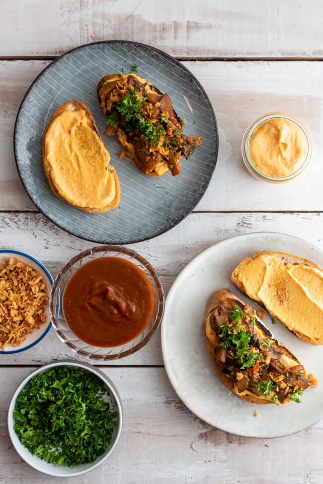 Veganes BBQ-Pilz-Sandwich auf grauen Keramiktellern mit BBQ-Sauce, Ranch-Sauce, Röstzwiebeln und frischen Kräutern dekoriert.