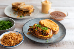 Veganes BBQ-Pilz-Sandwich auf grauen Keramiktellern mit BBQ-Sauce, Ranch-Sauce, Röstzwiebeln und frischen Kräutern dekoriert.