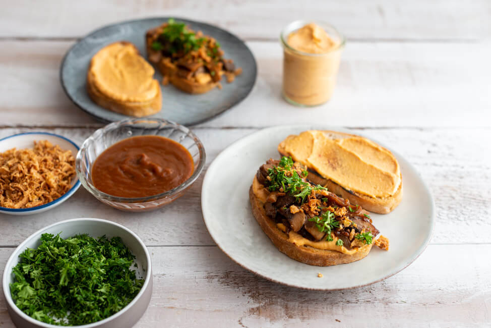 Veganes BBQ-Pilz-Sandwich auf grauen Keramiktellern mit BBQ-Sauce, Ranch-Sauce, Röstzwiebeln und frischen Kräutern dekoriert.