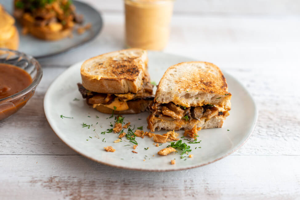Veganes BBQ-Pilz-Sandwich auf grauen Keramiktellern mit BBQ-Sauce, Ranch-Sauce, Röstzwiebeln und frischen Kräutern dekoriert.