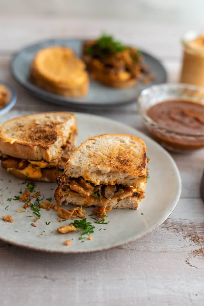 Veganes BBQ-Pilz-Sandwich auf grauen Keramiktellern mit BBQ-Sauce, Ranch-Sauce, Röstzwiebeln und frischen Kräutern dekoriert.