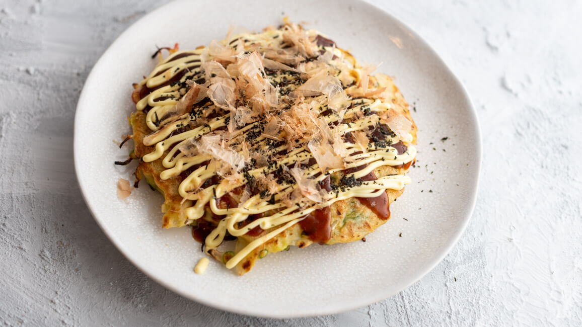 Originalrezept aus Kitchen Impossible: Okonomiyaki - japanischer Pfannkuchen - auf weißem Keramikteller. Garniert mit Okonomi Sauce, Kewpie, Aonori und Bonitoflocken.