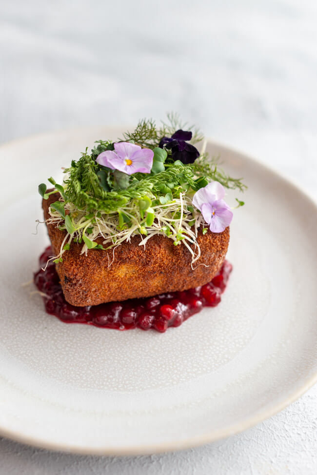 Rezept von Markus Stöckle aus Kitchen Impossible:: Cordon Bleu gefüllt mit Käsefondue und Bacon und belegt mit frischem Meerrettich, Wildkärutern und Blüten. Dazu Preiselbeeren und serviert auf einem weißen Teller.