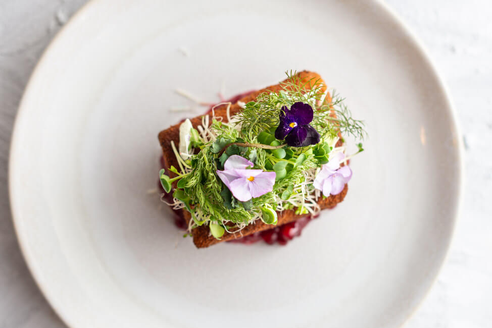 Rezept von Markus Stöckle aus Kitchen Impossible:: Cordon Bleu gefüllt mit Käsefondue und Bacon und belegt mit frischem Meerrettich, Wildkärutern und Blüten. Dazu Preiselbeeren und serviert auf einem weißen Teller.