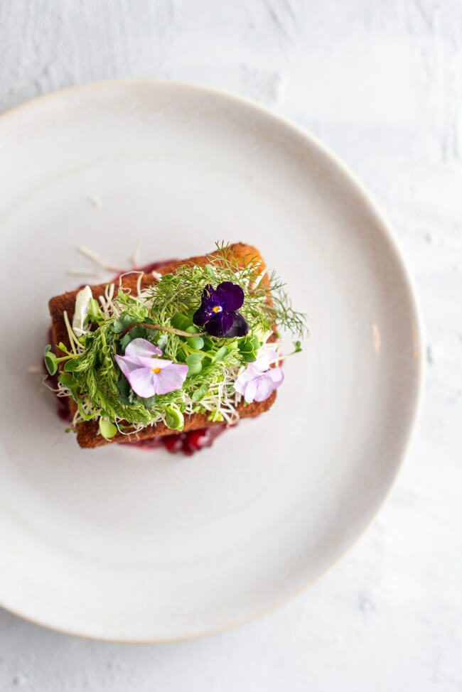 Rezept von Markus Stöckle aus Kitchen Impossible:: Cordon Bleu gefüllt mit Käsefondue und Bacon und belegt mit frischem Meerrettich, Wildkärutern und Blüten. Dazu Preiselbeeren und serviert auf einem weißen Teller.