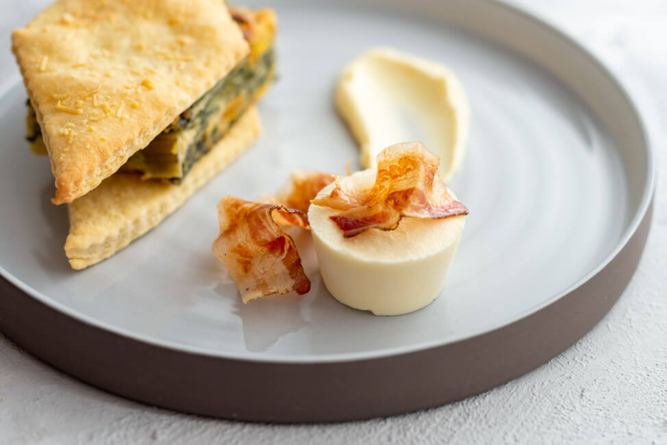 Erbazzone aus Kitchen Impossible: Mangold-Auflauf mit Blätterteig-Rauten, Parmesan-Eis mit knusprigem Panchetta und Parmesan-Creme auf hellem Keramikteller und hellem Untergrund.