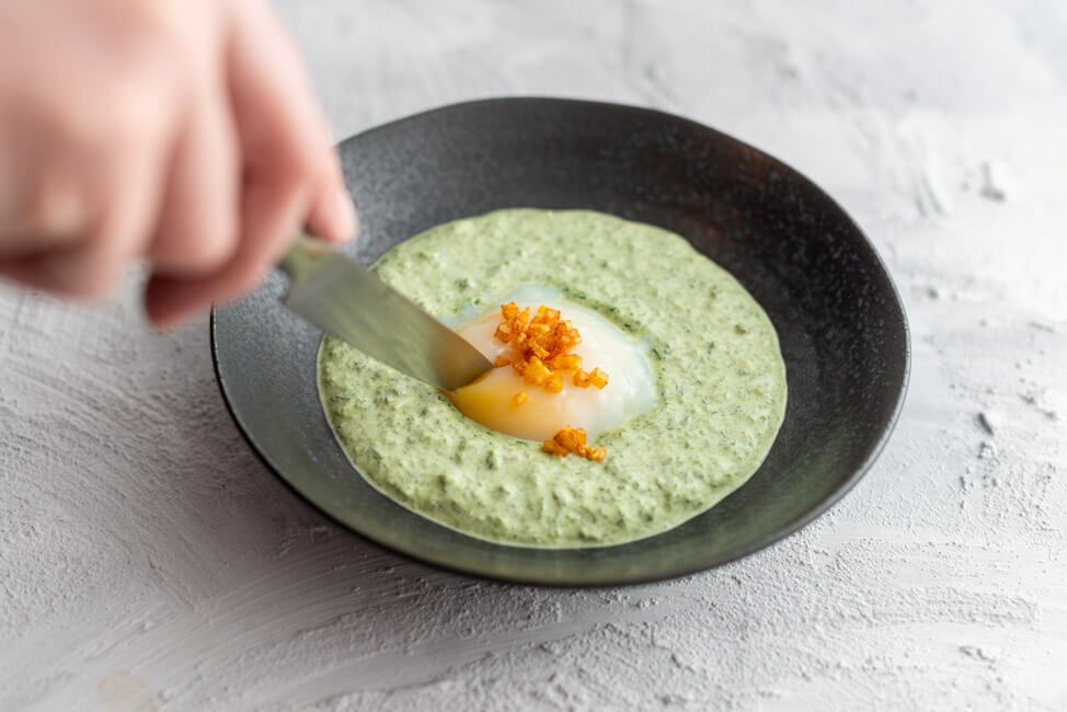 Rezept von Mario Lohninger aus Kitchen Impossible: Frankfurter Grüne Soße mit 60-Minuten-Ei und Kartoffel-Brunoises auf schwarzem Teller und hellem Untergrund.