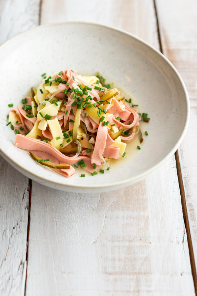 Allgäuer Wurstsalat wie auf der Hütte mit Käse und Gewürzgurken in weißer Porzellanschale auf weißem Holzuntergrund.