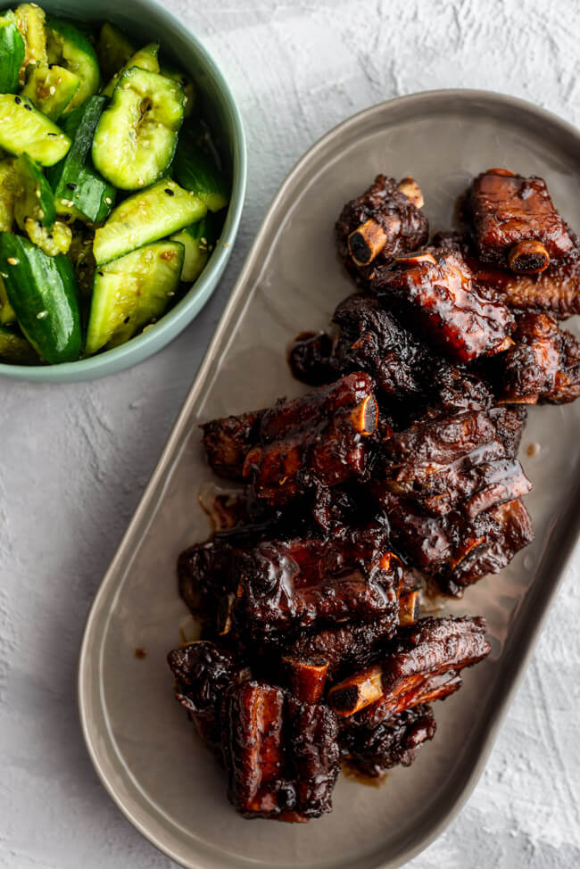 glasierte Chinesische Spare Ribs auf grauem Porzellanteller mit smashed cucumber salad in türkiser Porzellanschale auf hellem Untergrund.