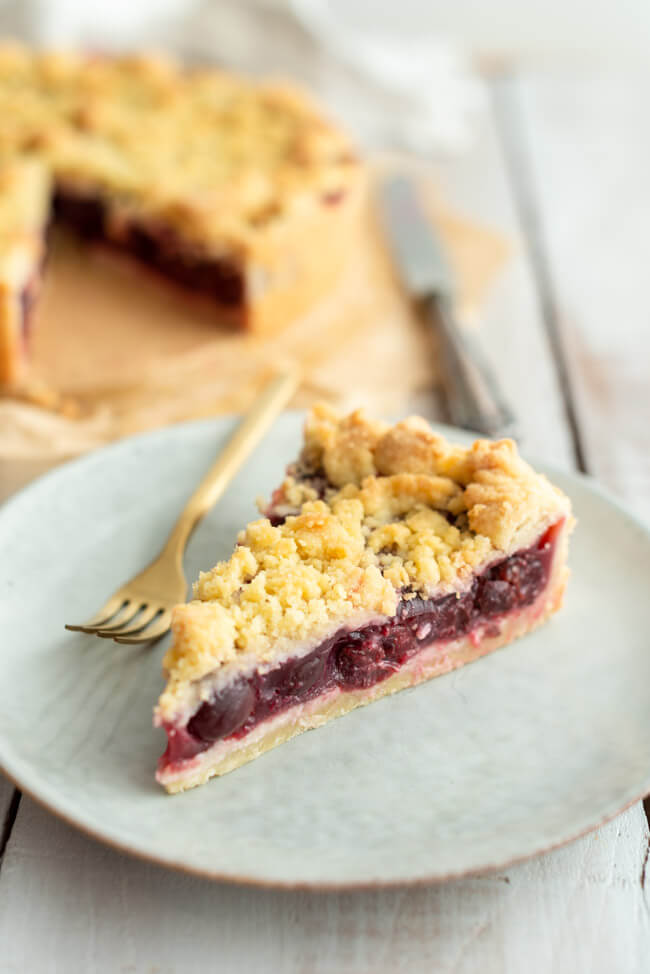 Ein Stück Kirsch-Streuselkuchen auf grauem Keramikteller und hellem Untergrund.