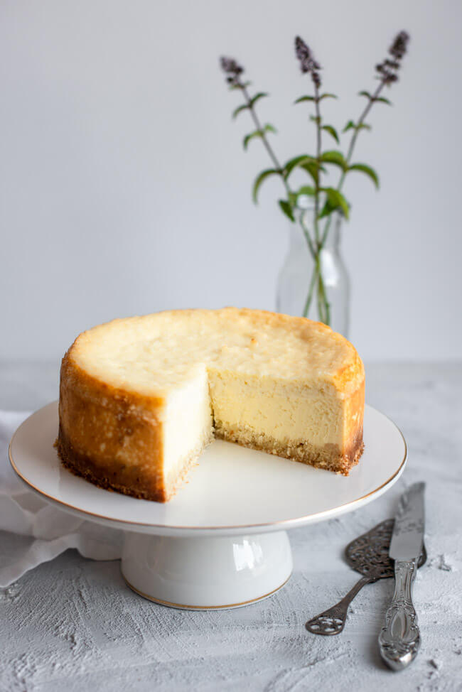 Kokos-Käsekuchen auf weißem Kuchenständer mit Goldrand und weißem Unter- und Hintergrund.