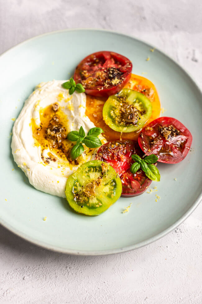 Labneh mit bunten Tomaten, Knoblauch-Gewürzöl und Basilikum auf blauem Keramikteller und hellem Untergrund.