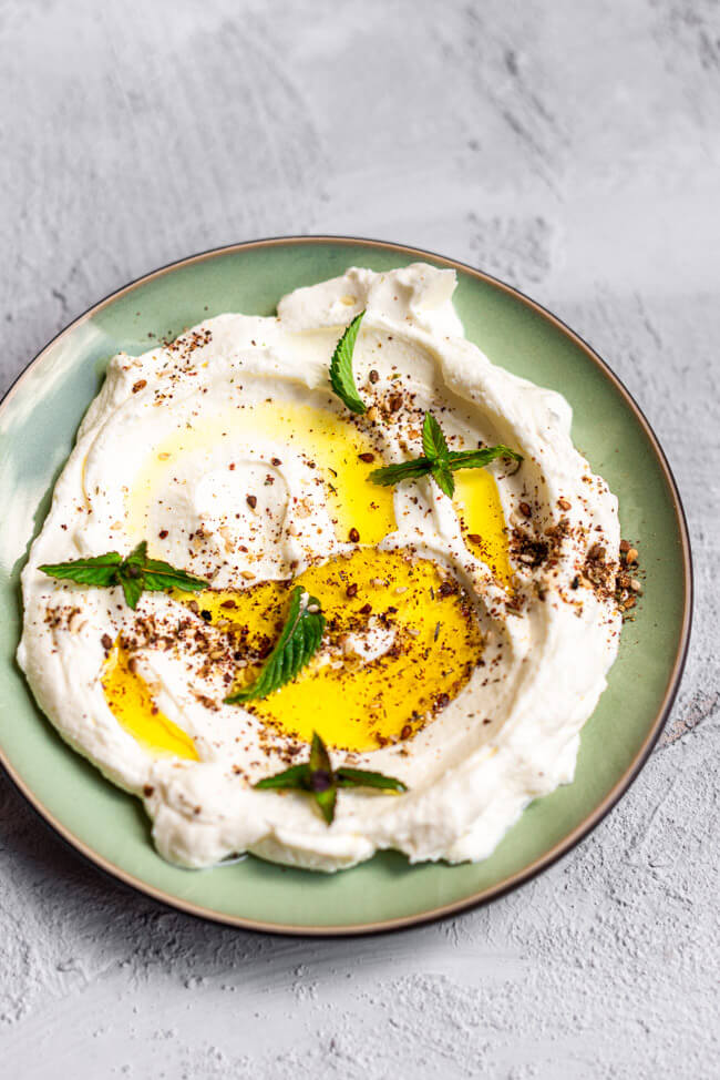 Labneh mit Olivenöl, Za'atar und frischer Minze auf grünem Teller und hellem Untergrund.