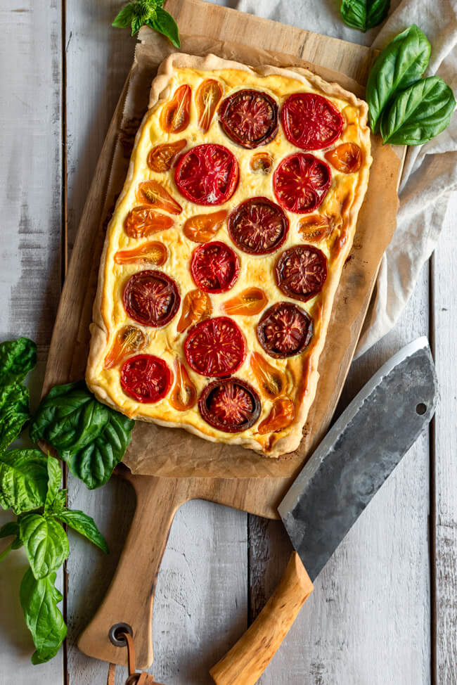 Tomaten-Tarte auf großem Holzbrett mit Basilikum und rustikalem Messer.
