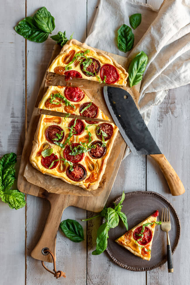 Tomaten-Tarte auf großem Holzbrett mit Basilikum und rustikalem Messer.