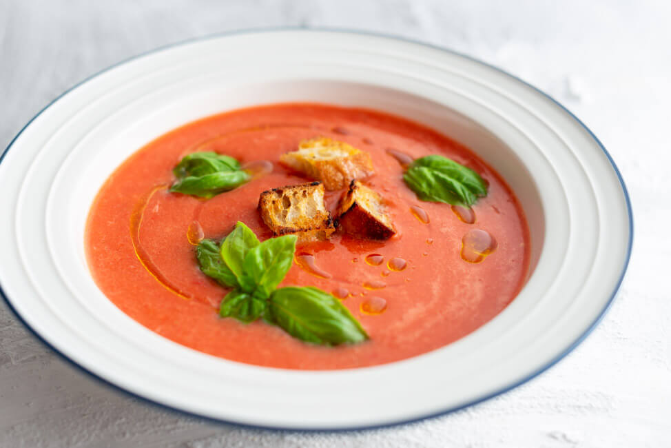 Wassermelonen Gazpacho mit Croutons und frischem Basilikum in weißem Teller mit blauem Rand.