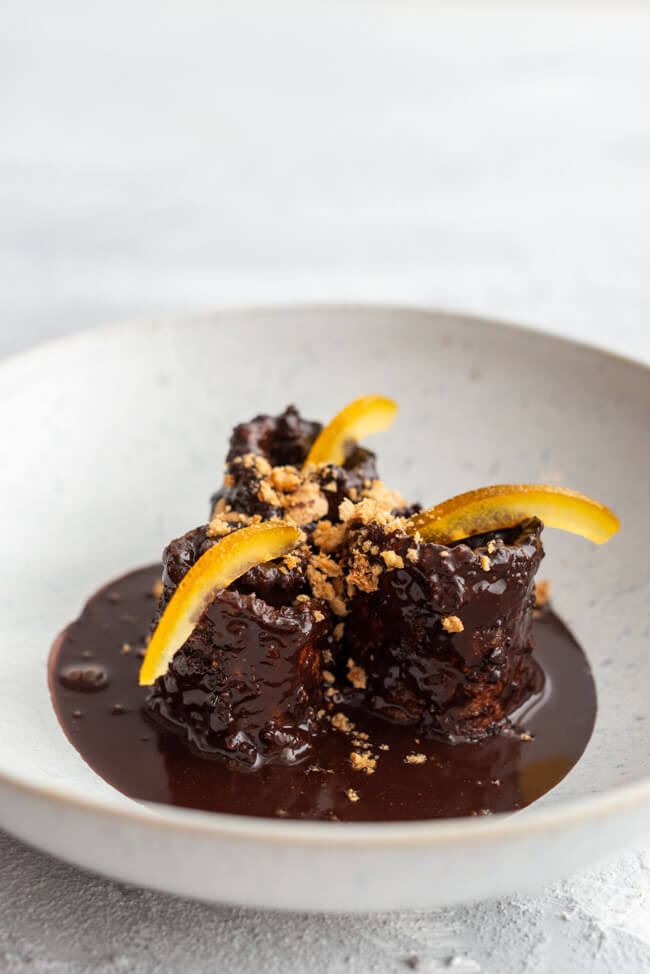 3 Auberginenröllchen mit Schokoladensauce, garniert mit kandierten Orangen und Amarettini auf weißem Untergrund.