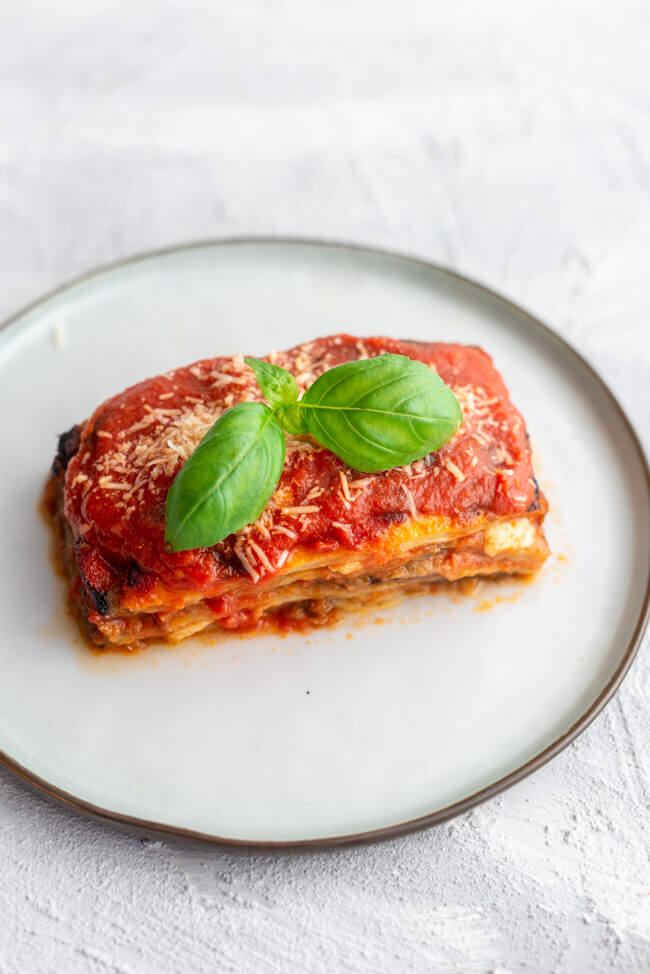 Ein Stück Melanzana alla Parmigiana, garniert mit Basilikum und Parmesan auf hellem Teller und weißem Untergrund.