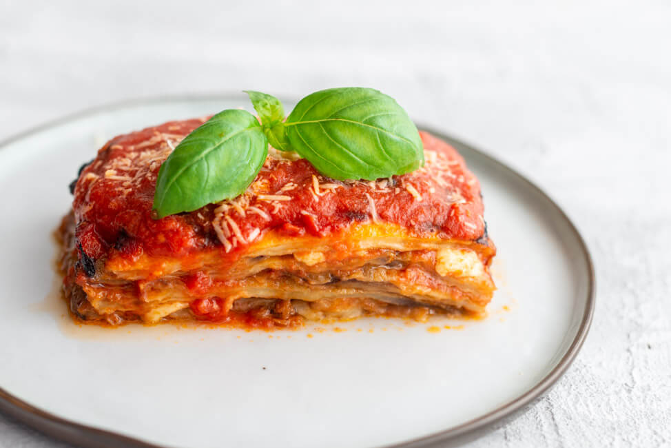 Ein Stück Melanzana alla Parmigiana, garniert mit Basilikum und Parmesan auf hellem Teller und weißem Untergrund.