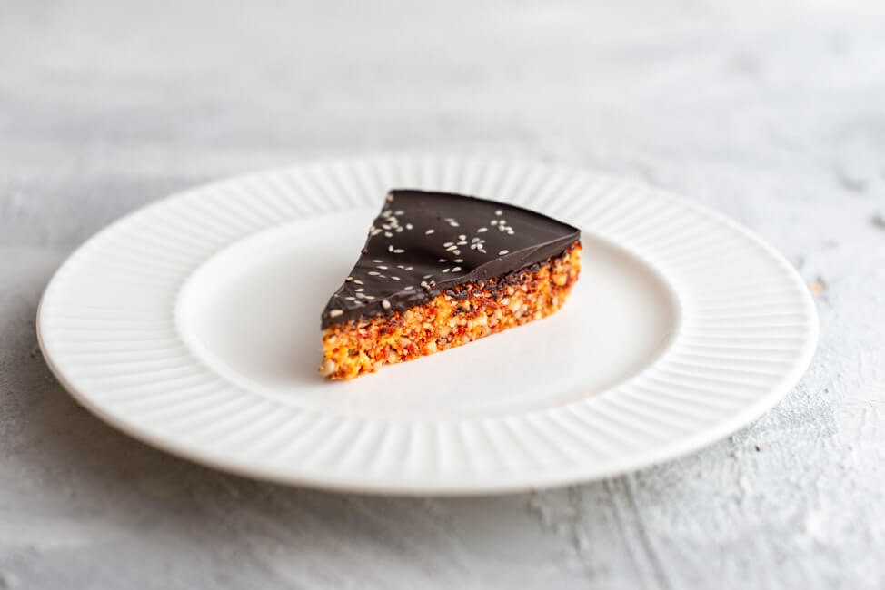 Ein Stück Power Cake, glasiert mit dunkler Schokolade, auf weißem Teller und hellem Untergrund.