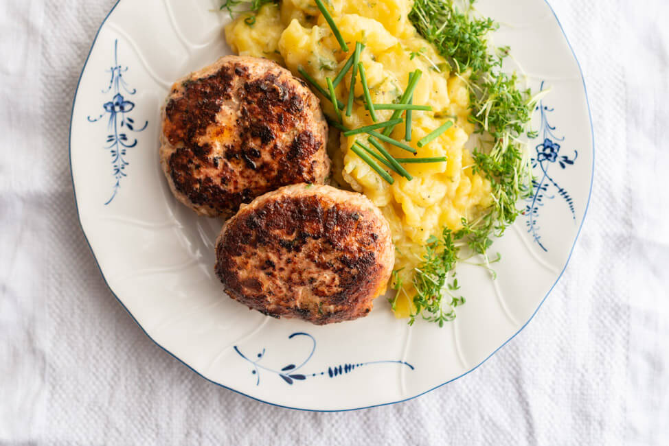 Zwei Fleischpflanzerl mit Kartoffelsalat garniert mit frischer Kresse und Schnittlauch auf weiß-blauem Porzellanteller auf weißer Tischdecke.