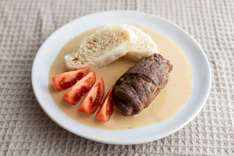 Roulade mit Böhmischen Knödeln und Tomatenschnitzen auf weißem Teller und beiger Tischdecke.