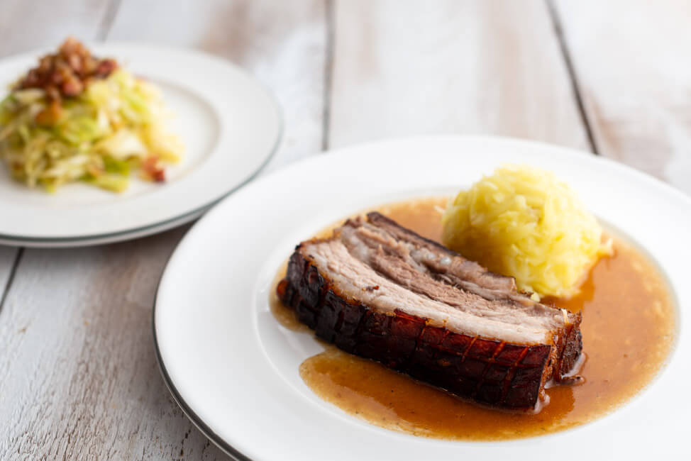 Eine Scheibe Krustenbraten mit Kloß und Soß und Krautsalat nach einem Rezept von Eckart Witzigmann auf weißem Porzellanteller und weißem Holztisch.