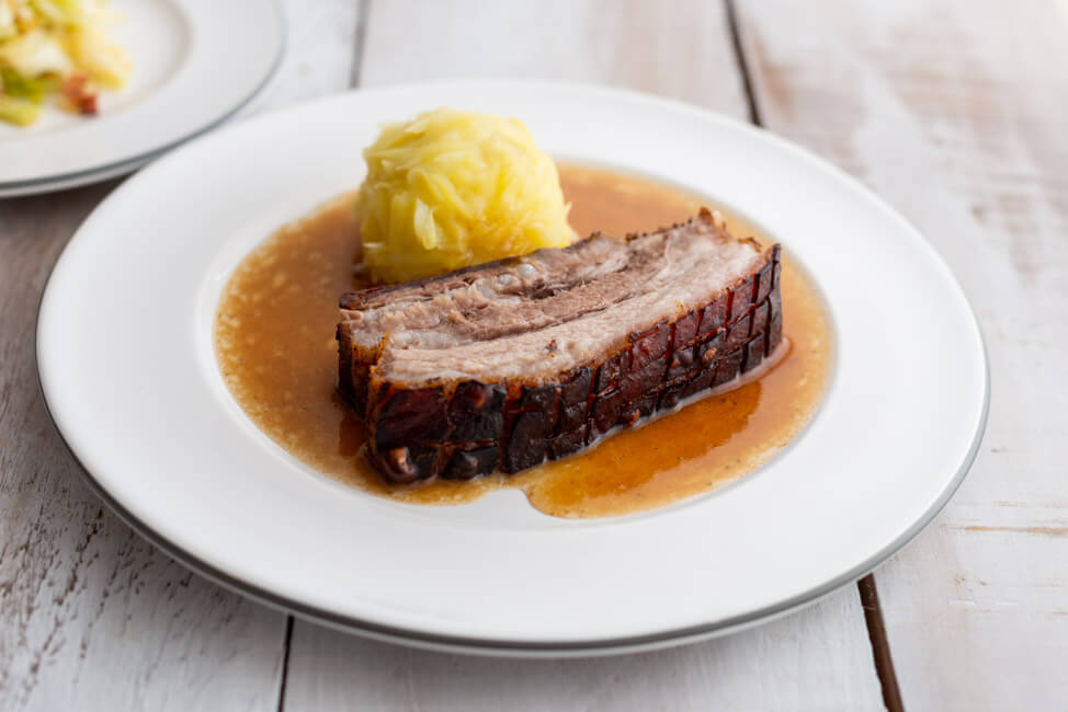 Eine Scheibe Krustenbraten mit Kloß und Soß und Krautsalat nach einem Rezept von Eckart Witzigmann auf weißem Porzellanteller und weißem Holztisch.