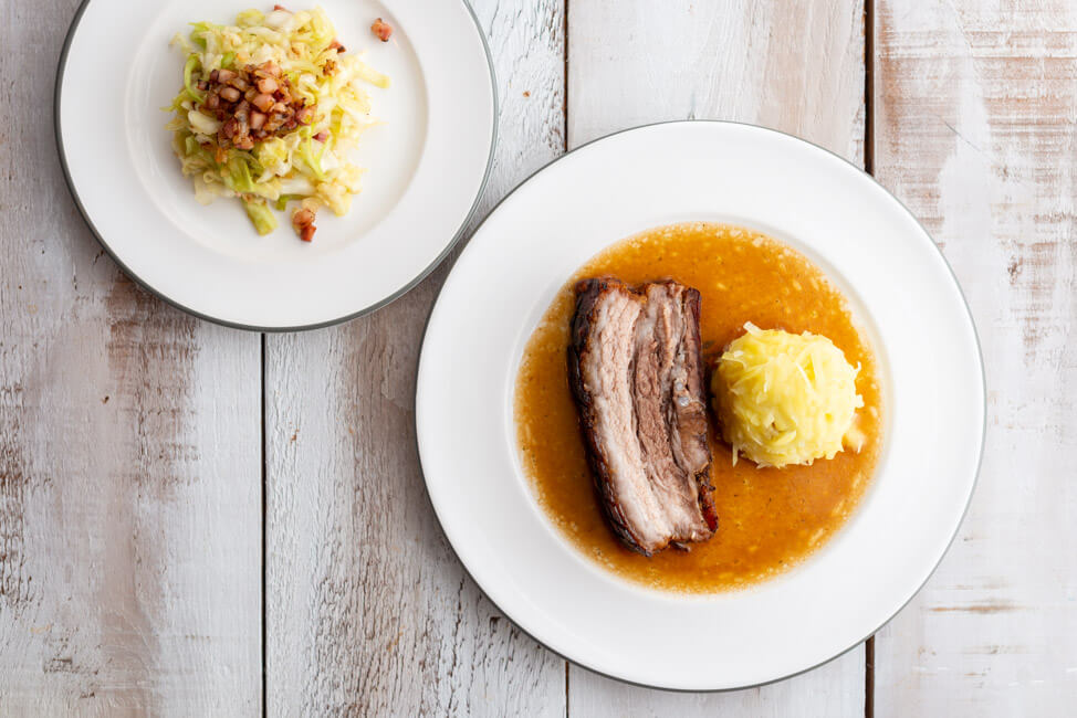 Eine Scheibe Krustenbraten mit Kloß und Soß und Krautsalat nach einem Rezept von Eckart Witzigmann auf weißem Porzellanteller und weißem Holztisch.