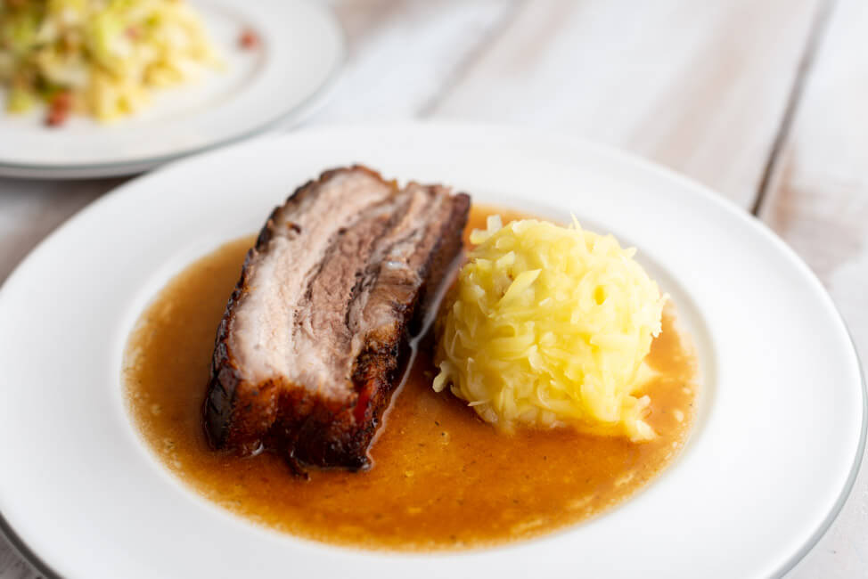 Eine Scheibe Krustenbraten mit Kloß und Soß und Krautsalat nach einem Rezept von Eckart Witzigmann auf weißem Porzellanteller und weißem Holztisch.