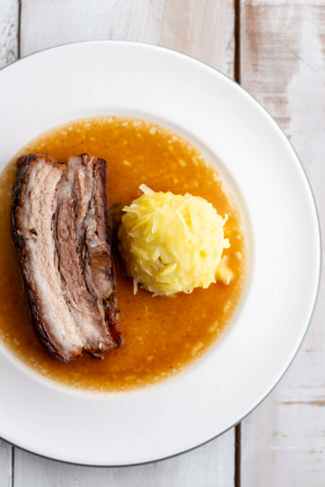 Eine Scheibe Krustenbraten mit Kloß und Soß nach einem Rezept von Eckart Witzigmann auf weißem Porzellanteller und weißem Holztisch.
