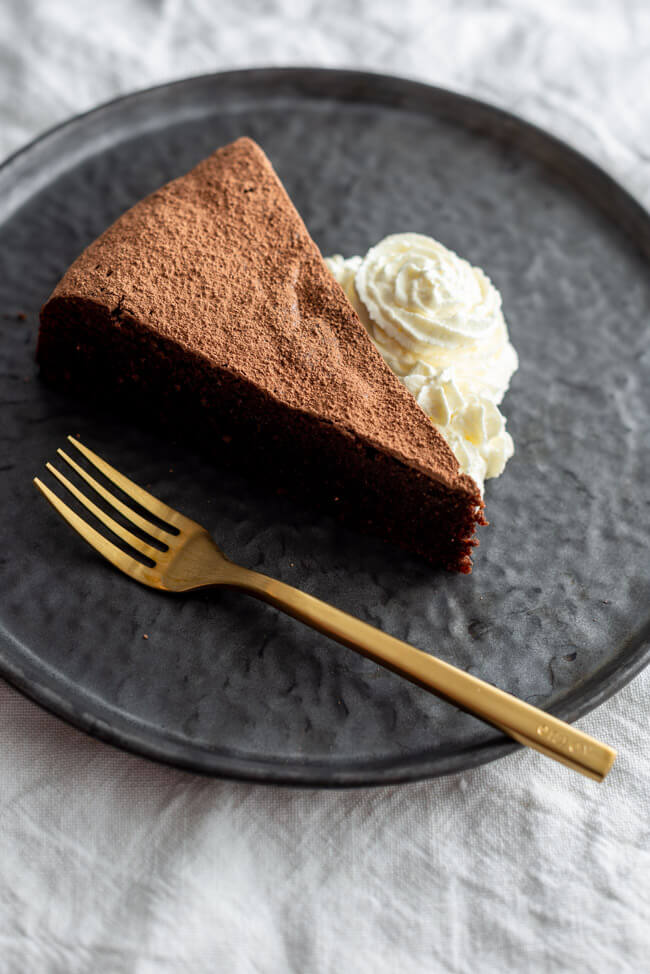 Ein Stück Schokoladenkuchen mit Sahne und goldener Gabel auf schwarzem Teller.