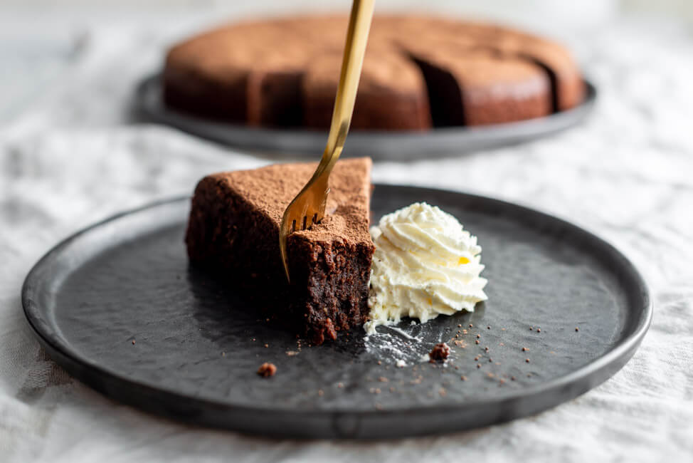 Ein Stück Schokoladenkuchen mit Sahne und goldener Gabel auf schwarzem Teller.