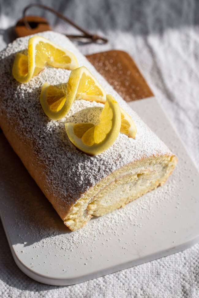 Feine Zitronen-Bisquitrolle auf Marmorbrett verziert mit Puderzucker und frischen Zitronenscheiben.