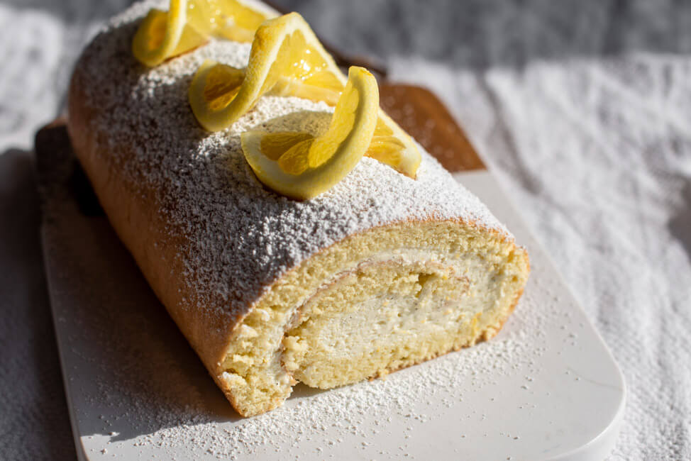 Feine Zitronen-Bisquitrolle auf Marmorbrett verziert mit Puderzucker und frischen Zitronenscheiben.