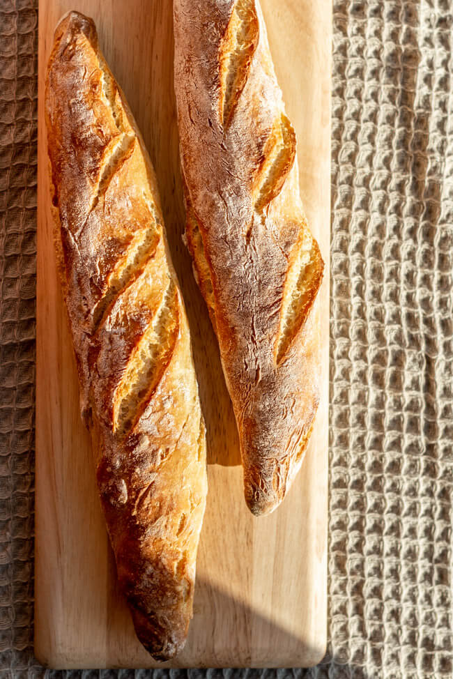 Zwei Baguette auf hellem Holzbrett und beige farbener Tischdecke im Sommenschein.