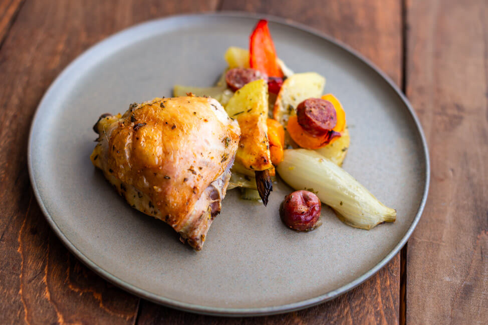 Hühnerteile, Gemüse und Salsiccia auf grünem Keramikteller und dunklem Holztisch.