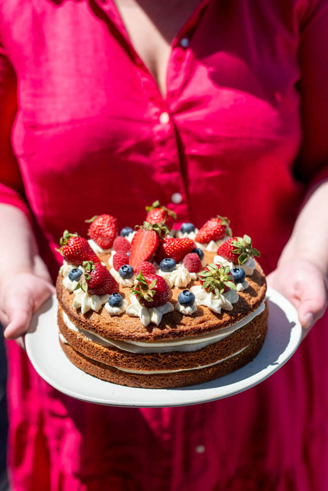 Frau in rotem Kleid hält in ihren Händen einen Teller auf dem die Sommerliche Beerentorte steht.