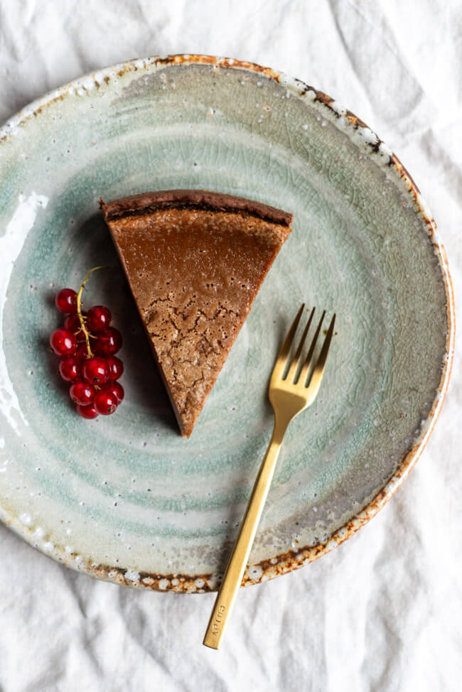 Ein Stück Nougatkäsekuchen mit frischen Johannisbeeren und goldener Gabel auf hellgrünem Keramikteller.