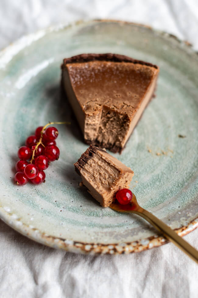 Ein Stück Nougatkäsekuchen mit frischen Johannisbeeren und goldener Gabel auf hellgrünem Keramikteller.