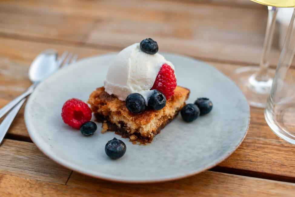 Ein Stück Pouding Chomeur mit frischen Beeren und einer Jugel Joghurteis auf Porzellanteller und Holztisch.