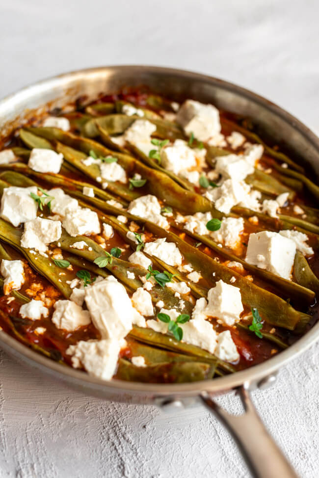 Geschmorte Stangenbohnen in Tomatensauce mit Feta in Kupferpfanne auf hellem Untergrund.