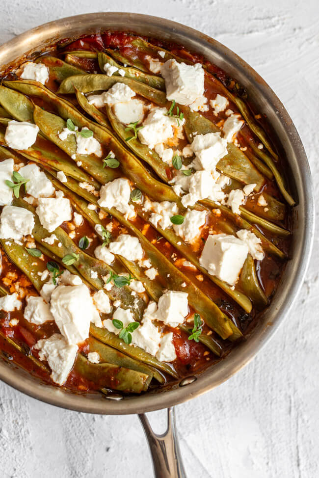 Geschmorte Stangenbohnen in Tomatensauce mit Feta in Kupferpfanne auf hellem Untergrund.